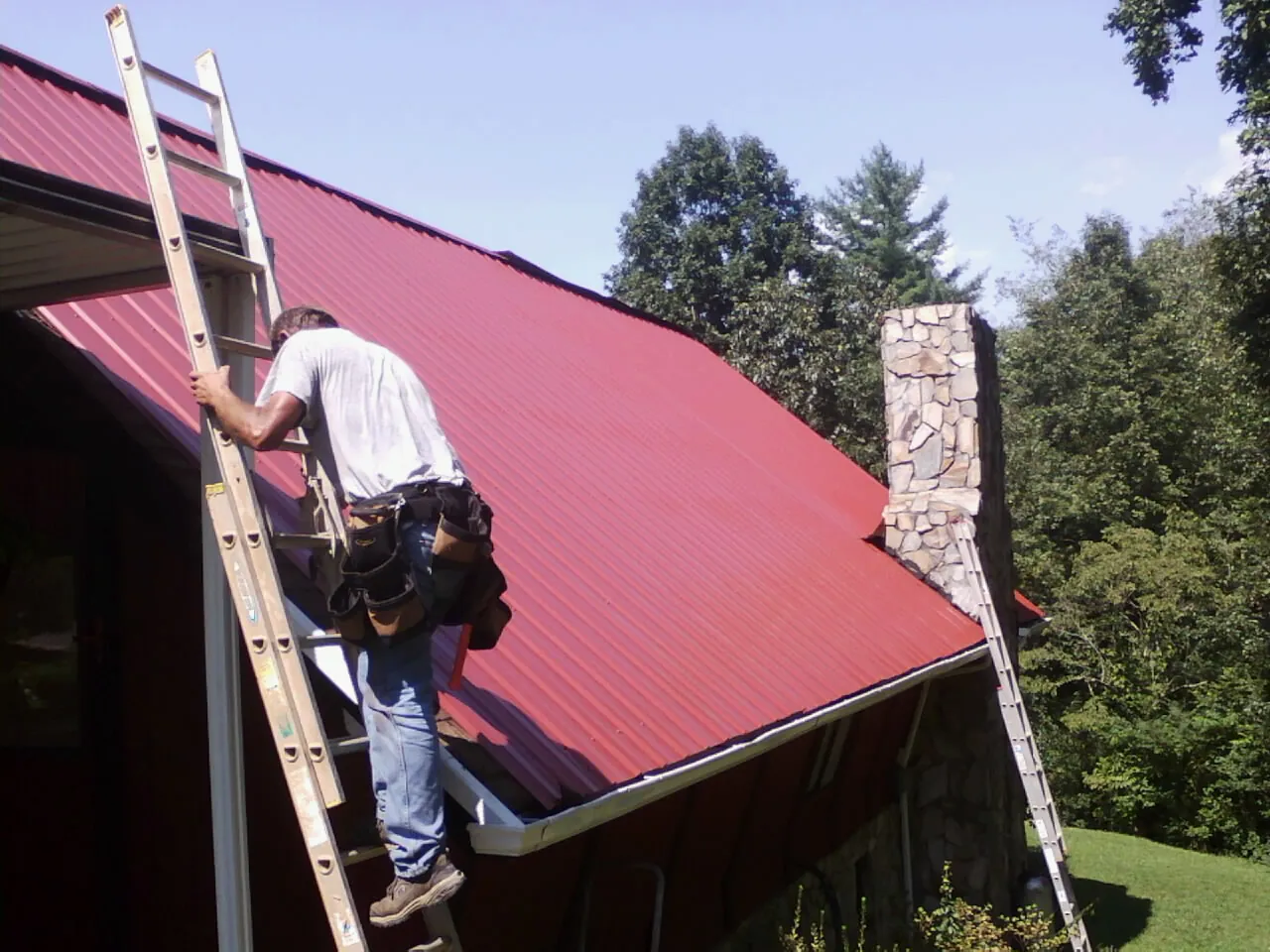 Roofer on a ladder beside a red metal roof and stone chimney — Soffit & Fascia Repair by Roofing Brothers in Sylvester